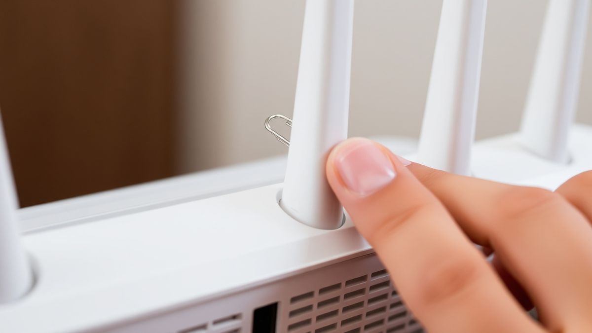 a person using a paperclip to press the reset button on the back of a router, with LED lights blinking